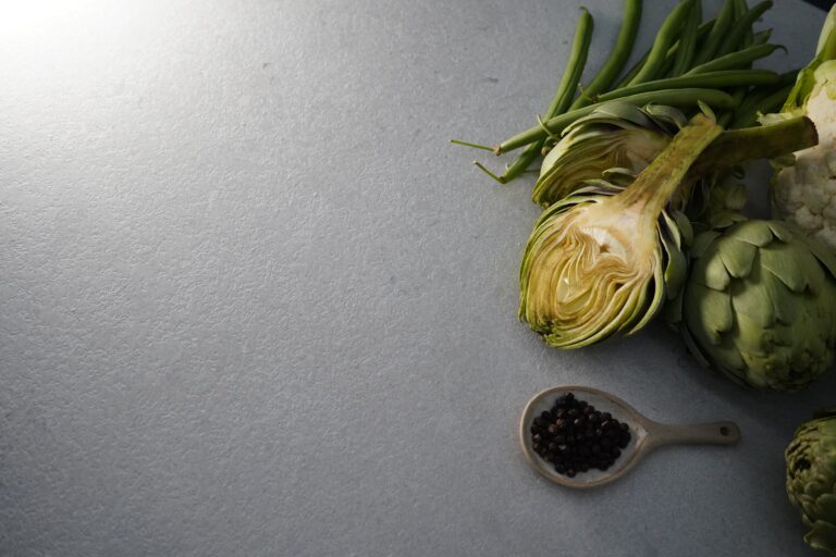 Superficie de piedra sinterizada Portland Pearl en primer plano, con fondo nítido que muestra vegetales frescos como alcachofas, coliflor y granos de pimienta negra, en estilo bodegón.