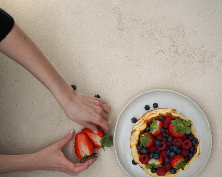 Manos colocando fresas junto a tarta de queso decorada con frutas del bosque sobre una superficie de piedra sinterizada de tono cálido.