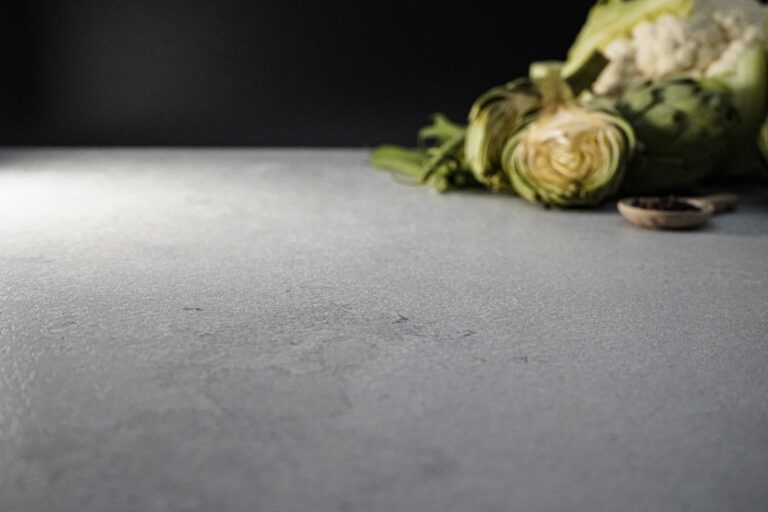 Superficie de piedra sinterizada Portland Pearl en primer plano, con un fondo desenfocado que muestra vegetales frescos como alcachofas y coliflor, estilo bodegón.